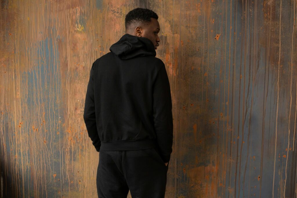 Back view of unrecognizable African American male wearing trendy black outfit standing near wooden wall with hands in pockets in room