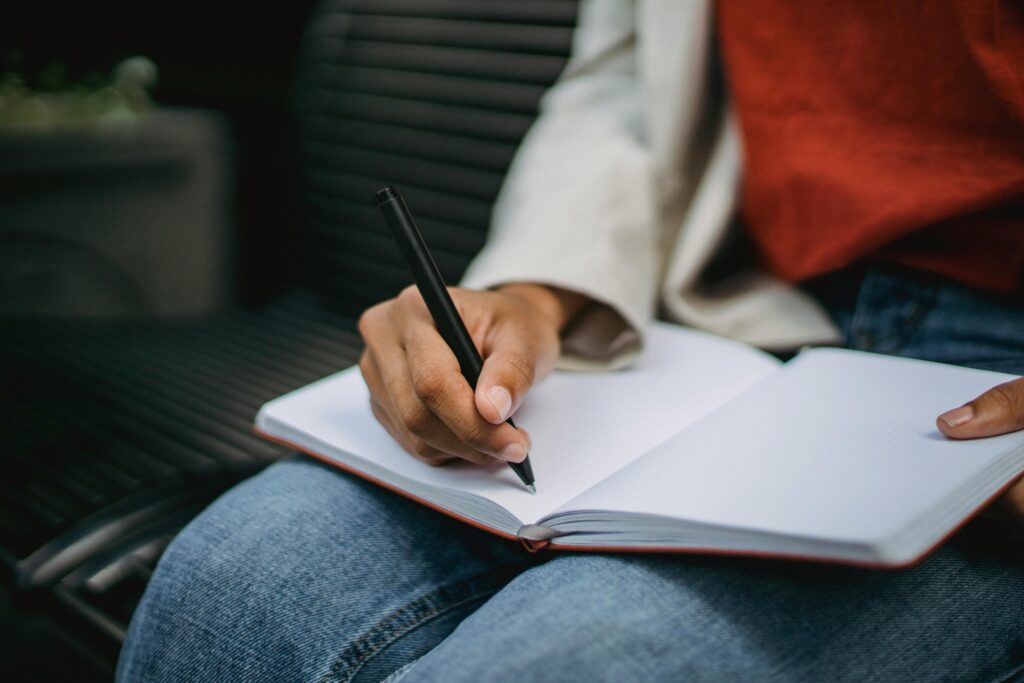 Adult writing notes in a notebook while seated outdoors, pen in hand.